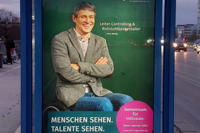 Werbeplakat an einer Bushaltestelle mit Leiter Controlling und Rollstuhlbasketballer Arne Wettig, Menschen sehen. Talente sehen. Gemeinsam für Inklusion, deine Spende hilft! www.pfennigparade.de