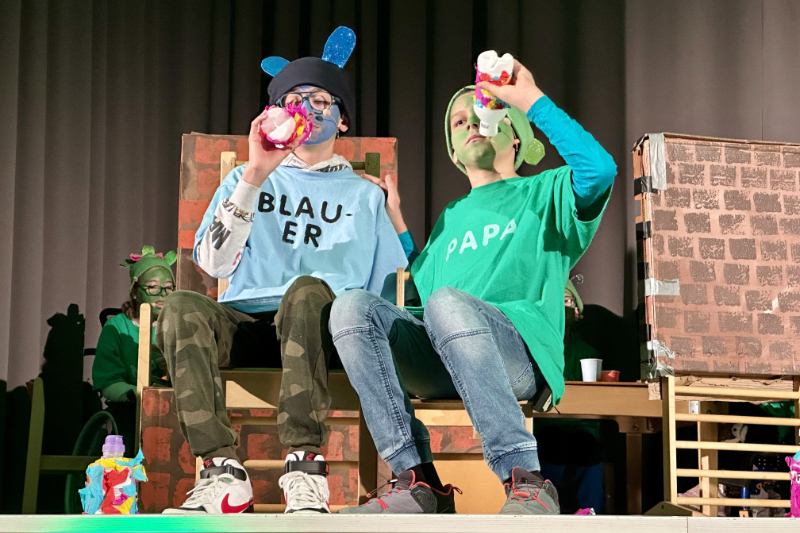 Zwei Schüler*innen verkleidet neben Theaterrequisiten auf der Bühne mit Sprossenstuhl
