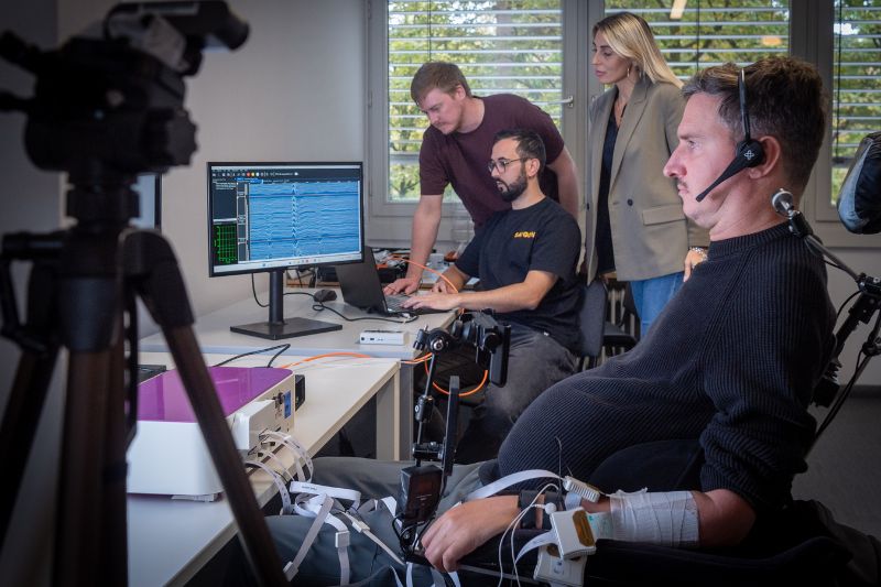 Rollstuhlfahrer mit Elektroden an den Armen und drei Wissenschaftler*innen schauen auf Computerbildschirm