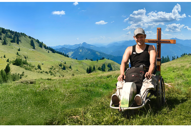 Mann mit Rollstuhl am Gipfelkreuz in Berglandschaft