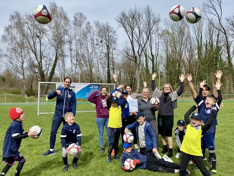 Kinder, Erwachsene und Jugendliche auf einem Fußballplatz werfen Bälle nach oben.