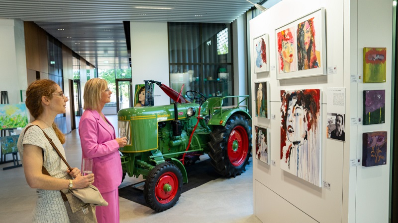 Zwei weibliche Personen betrachten Gemälde, im Hintergrund steht ein Traktor.