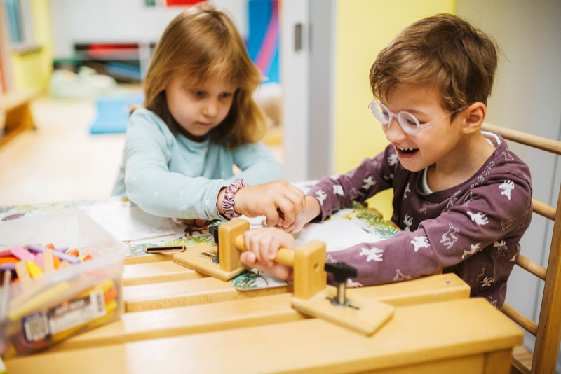 Zwei Kinder spielen an einem Tisch. Ein Kind sitzt auf einem Sprossenstuhl