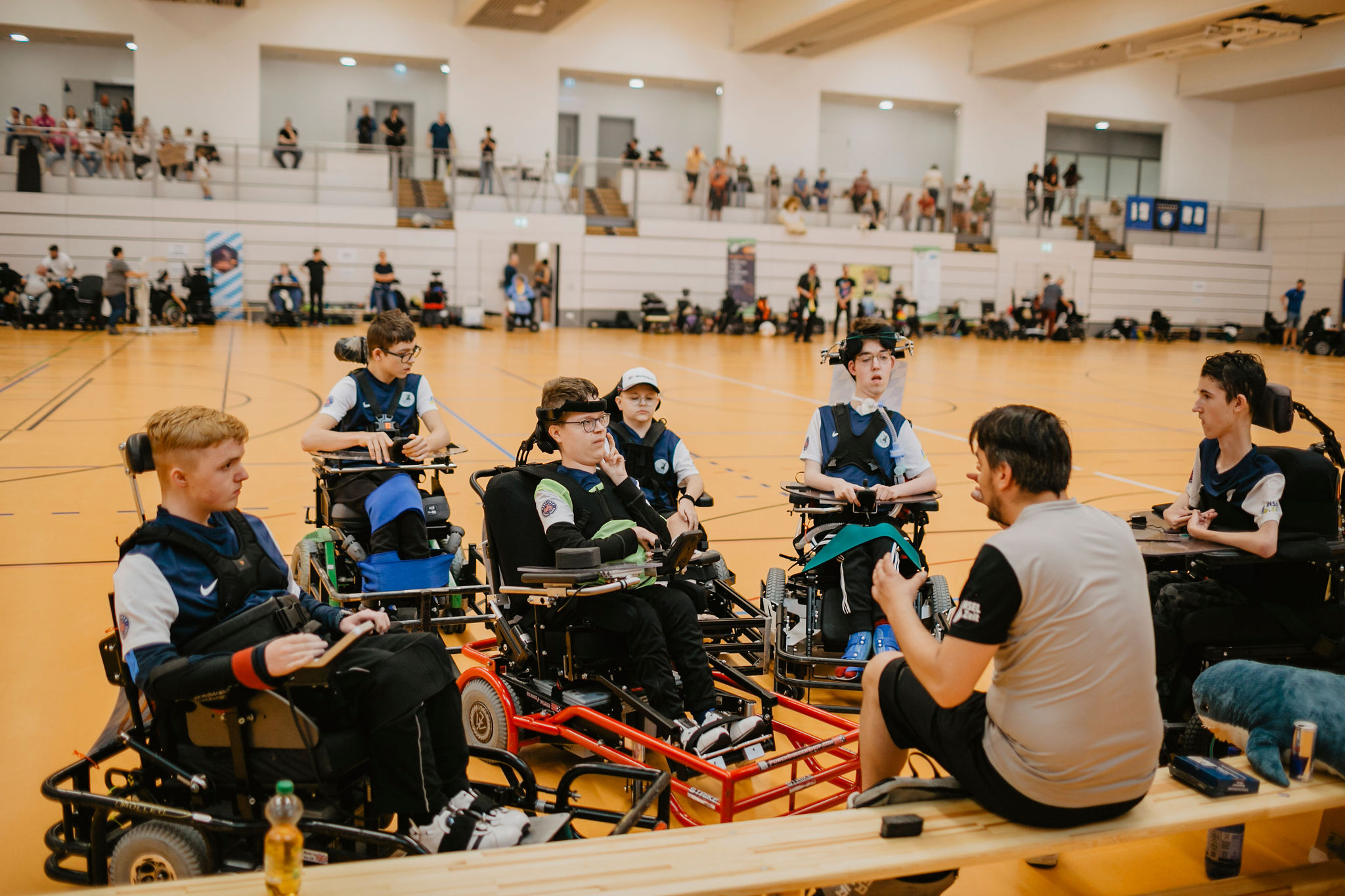 Sieben Spieler im E-Rollstuhl der Mannschaft "Munich Sharks" im Gespräch mit dem Trainer, der auf einer Bank im Vordergrund sitzt.