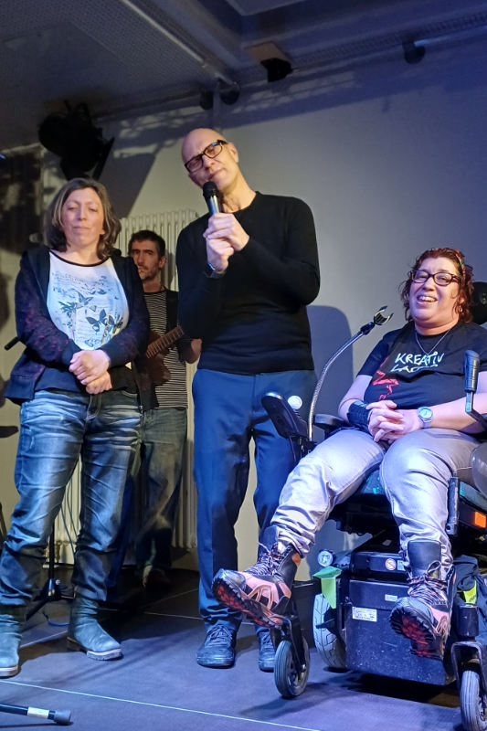 Drei Personen mit und ohne Rollstuhl auf der Bühne. Mann mit Mikrofon