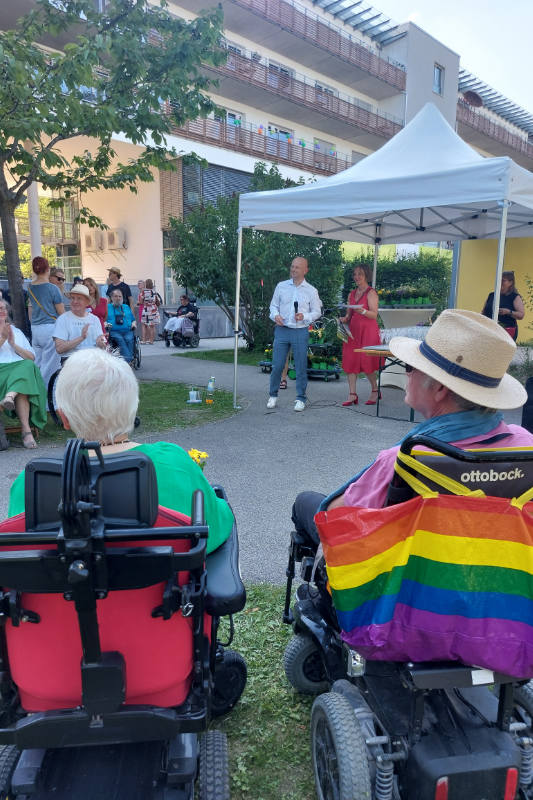 Zwei Personen stehen im Pavillion , mehrere Personen mit und ohne Rollstuhl davor auf der Wiese