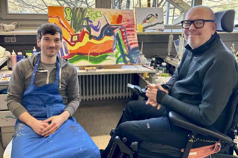 Zwei Künstler vor Kunstwerken. Einer davon im Rollstuhl