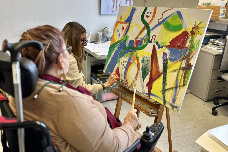 Zwei Personen vor Bild auf Staffelei mit Pinsel in der Hand.