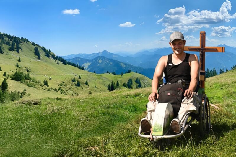 Person im Rollstuhl auf dem Berg neben einem Gipfelkreuz