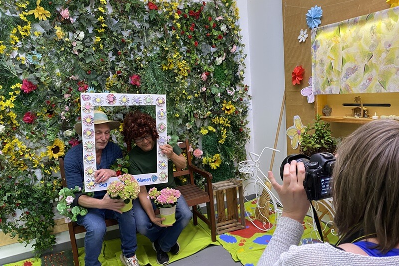 Zwei Personen schauen durch einen Bilderrahmen und sitzen vor einer Blumenwand.