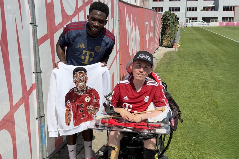 FC Bayern Fußballspieler Alonso Davies hält einen Pullover mit seinem Gesicht, daneben Danny im Rollstuhl