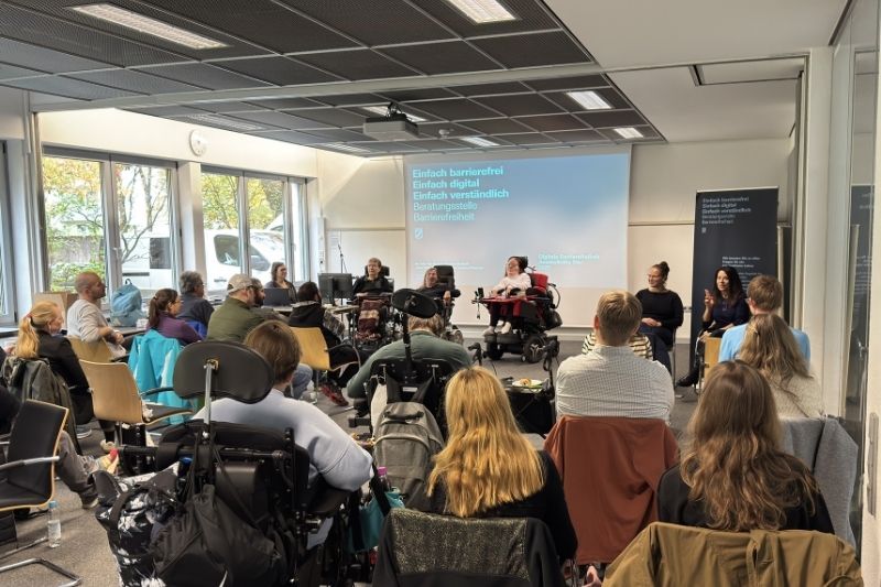 Vortragsituation. Gruppe von Menschen schaut in Richtung einer Präsentation, die von Personen im Rollstuhl gehalten wird.
